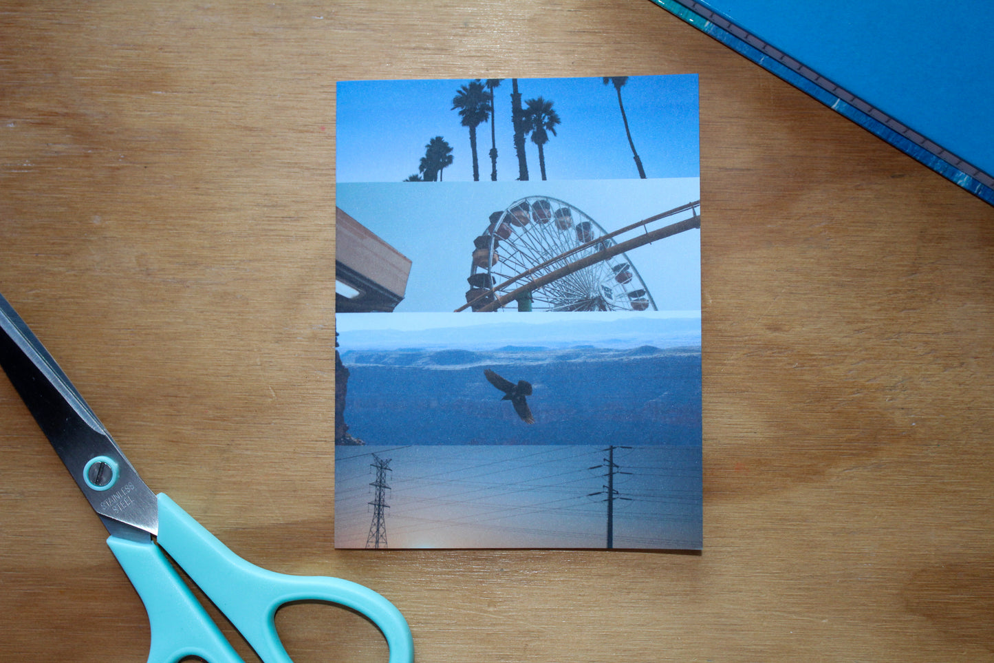 blue monochrome photo card with notebook and blue scissors in the picture - palm trees, ferris wheel, raven, and electric lines - on wood background