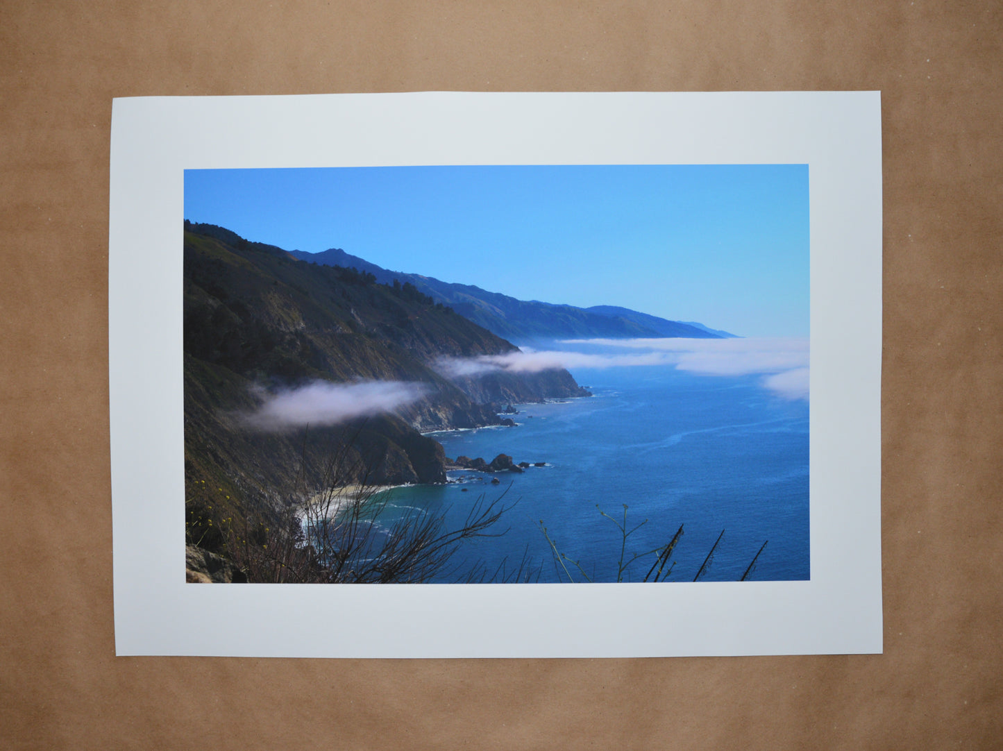 2 in borders with centered print of Big Sur coast line view of the moutains meeting the water. Deep blue hues with the marine layer of clouds suspended halfway of the mountain height. Rocks along the coast with blue skies. Plants in the foreground.
Kraft paper background.