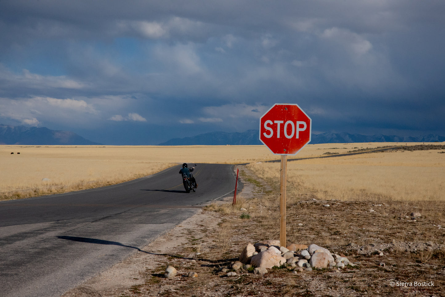 skyline with mountains, bright red stop sign with rocks at the base of the post in the foreground. the road with a man on a motorcycle.