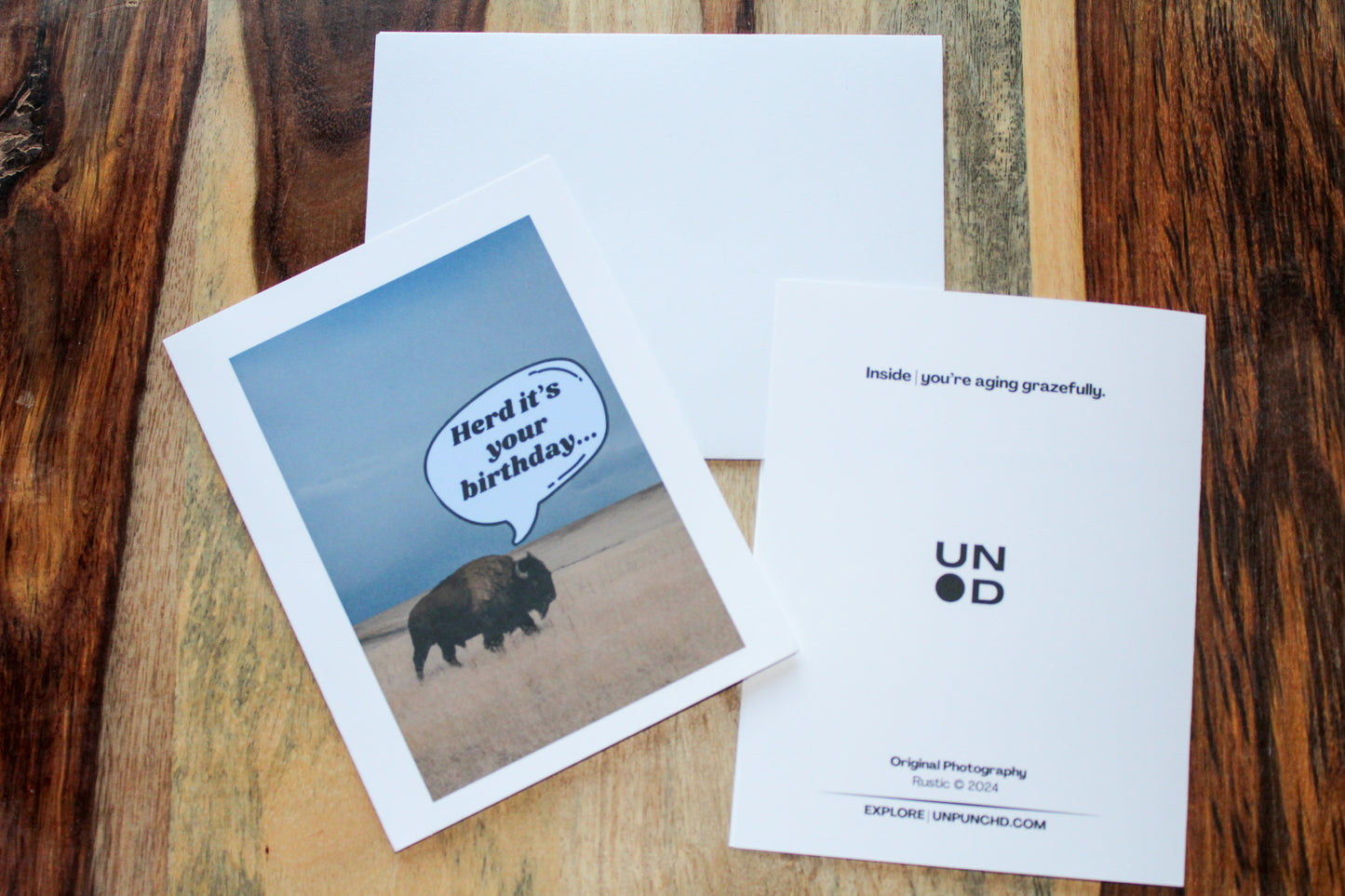 Card front and open card behind on stained wood. "herd it's your birthday..." American bison on the front grazing through a field with a blue sky. "Inside | you're aging grazefully." on the back with logo at the center card and original photography with Rustic, 2024 and the website URL at the bottom.