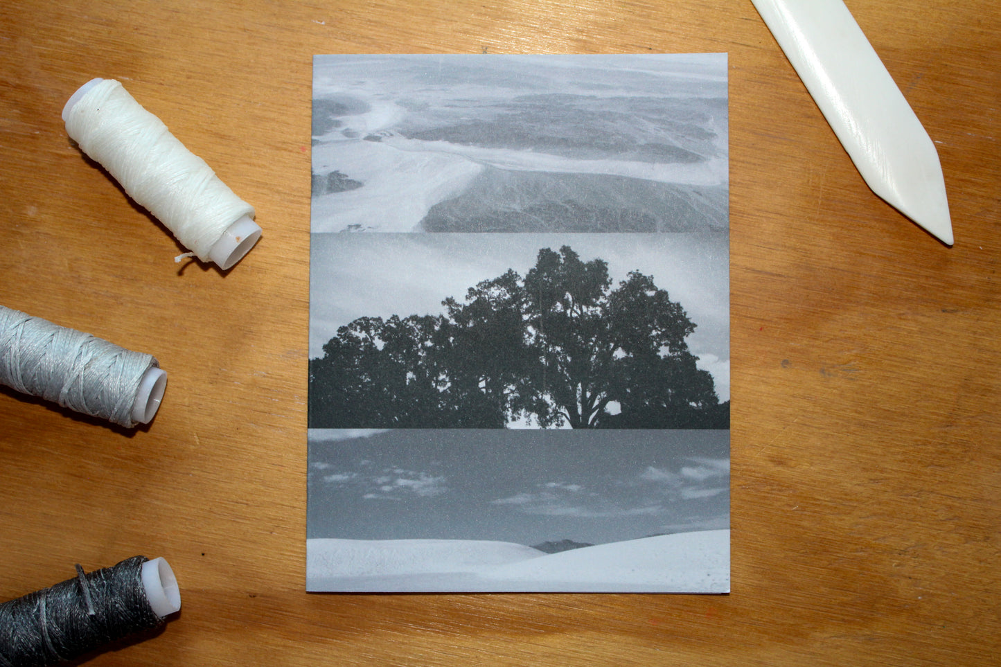 3 greyscale photos - desert, tree, and white sands. with a scoring tool and 3 grey wax linen thread spools. On a wood surface.