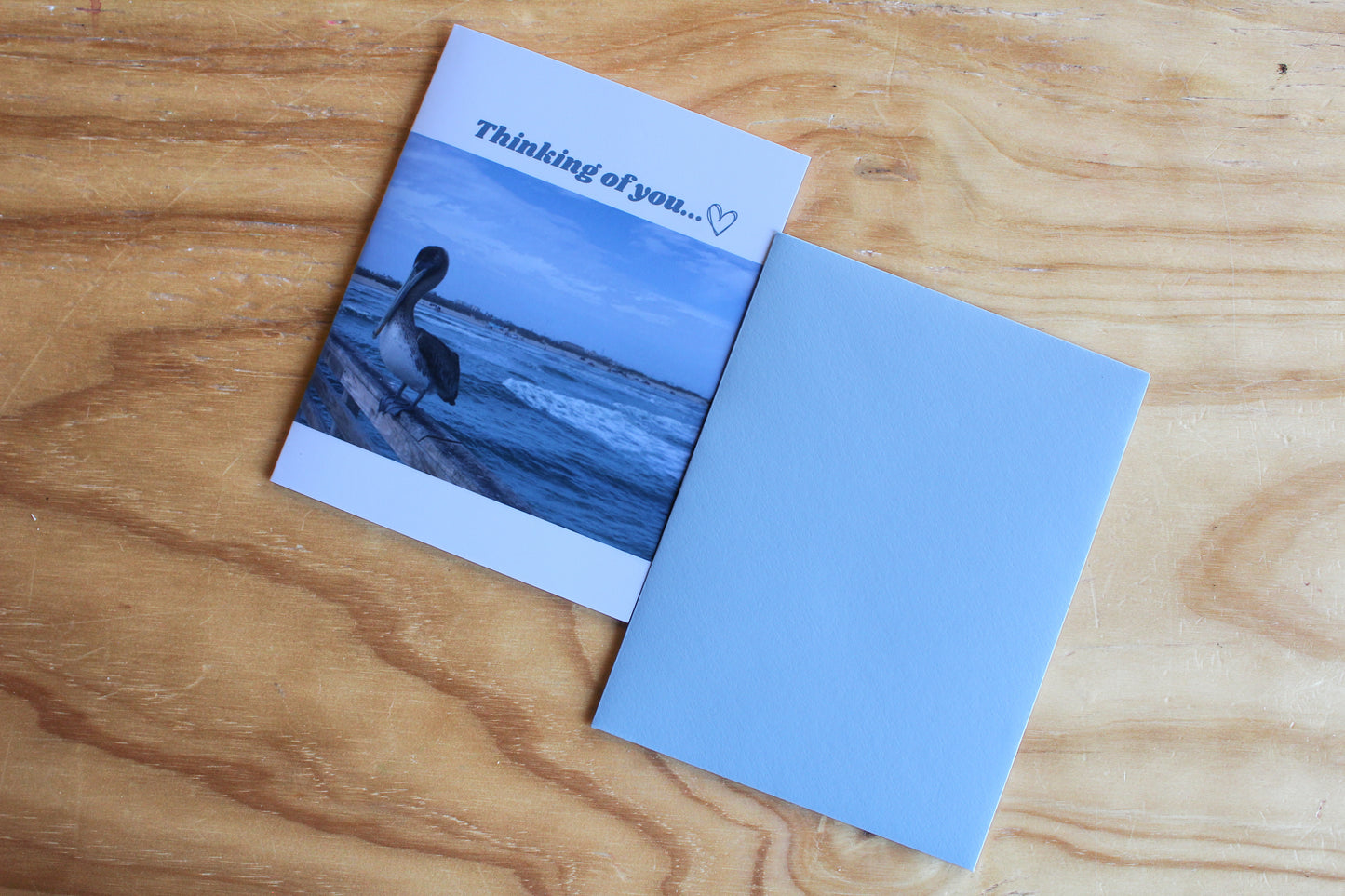 Thinking of you across the top with a delicate heart. Image mid card with a perched pelican against an oceanic background. Light blue envelope on a wood background.