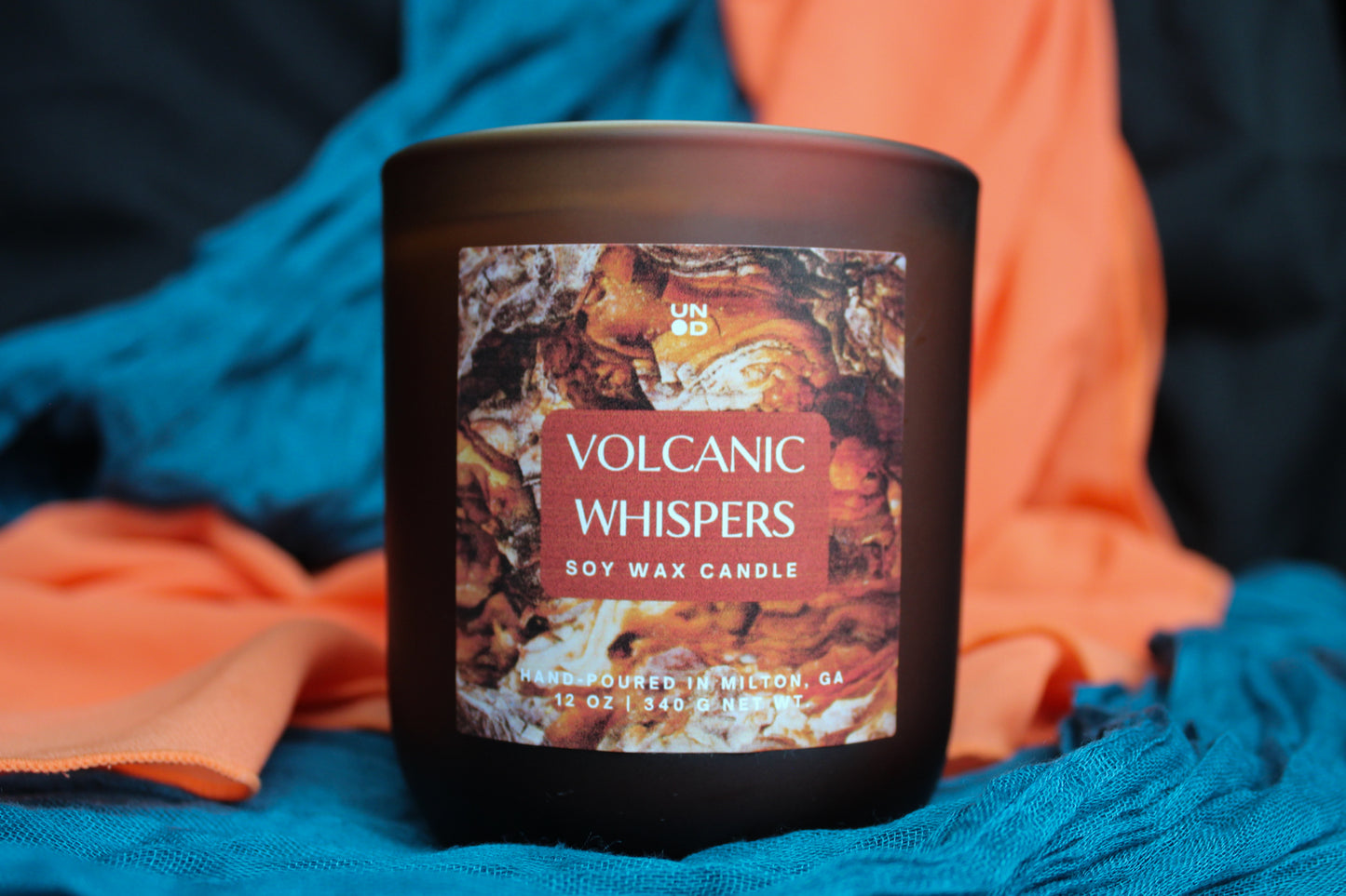 Soy wax candle labeled 'Volcanic Whispers' on a blue and orange fabric background