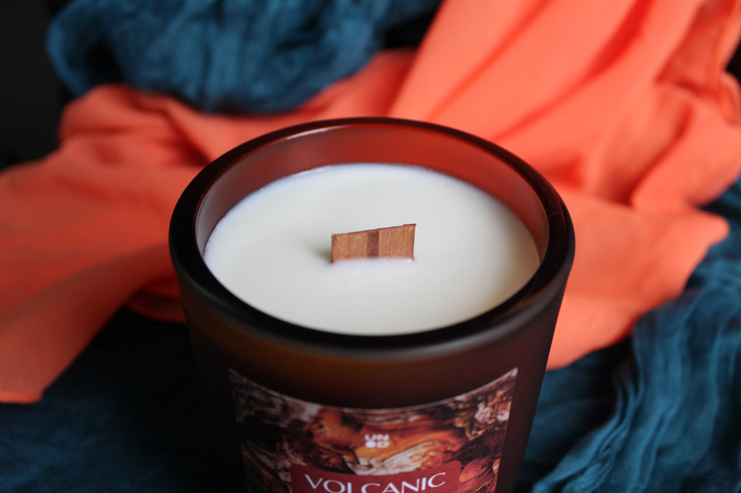 Candle labeled 'Volcanic' with a wooden wick, surrounded by orange and blue fabric.