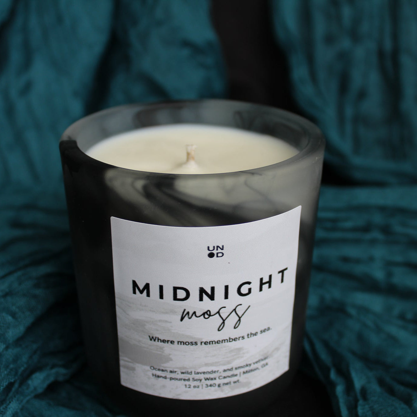 Candle labeled 'Midnight Moss' on a textured teal fabric background