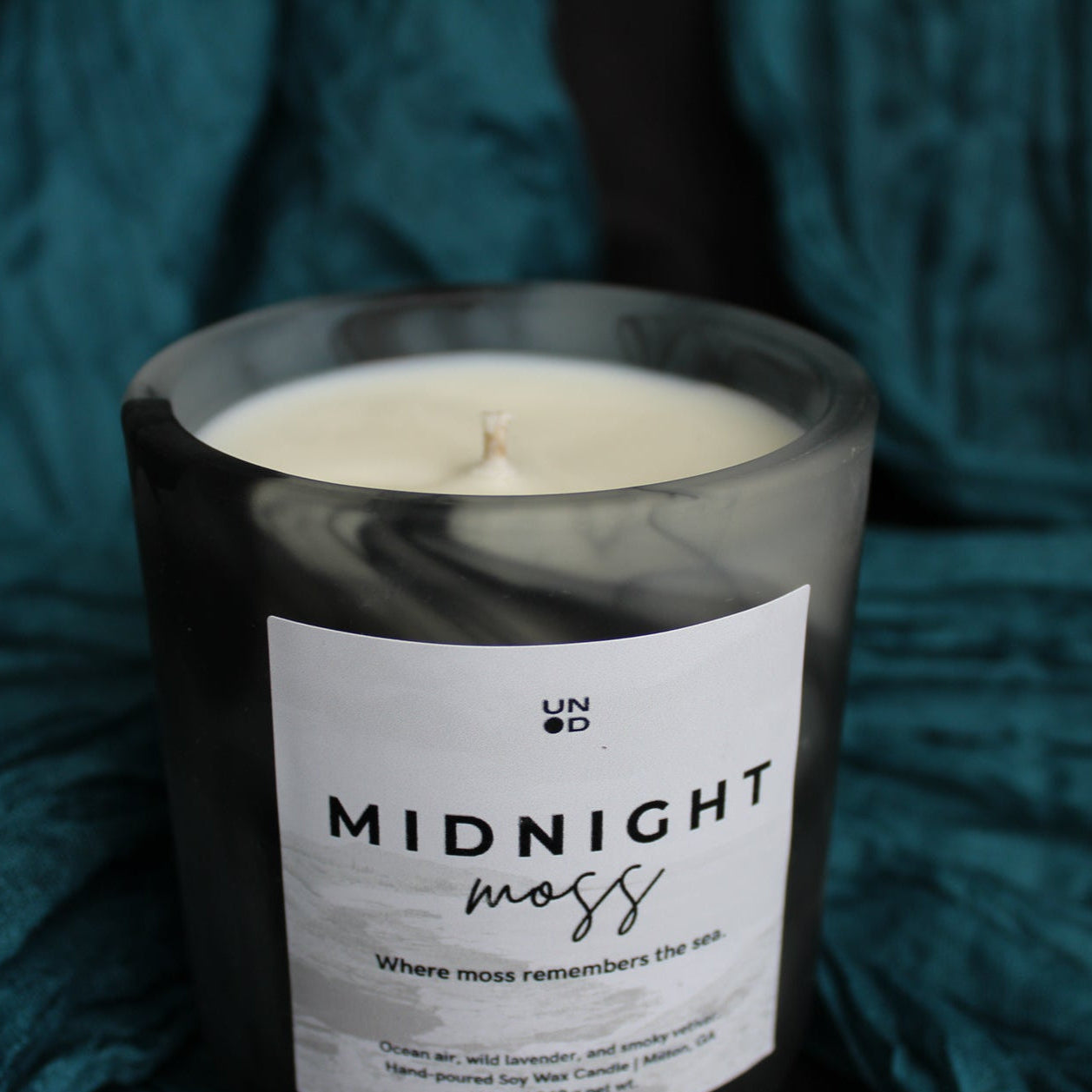 Candle labeled 'Midnight Moss' on a textured teal fabric background