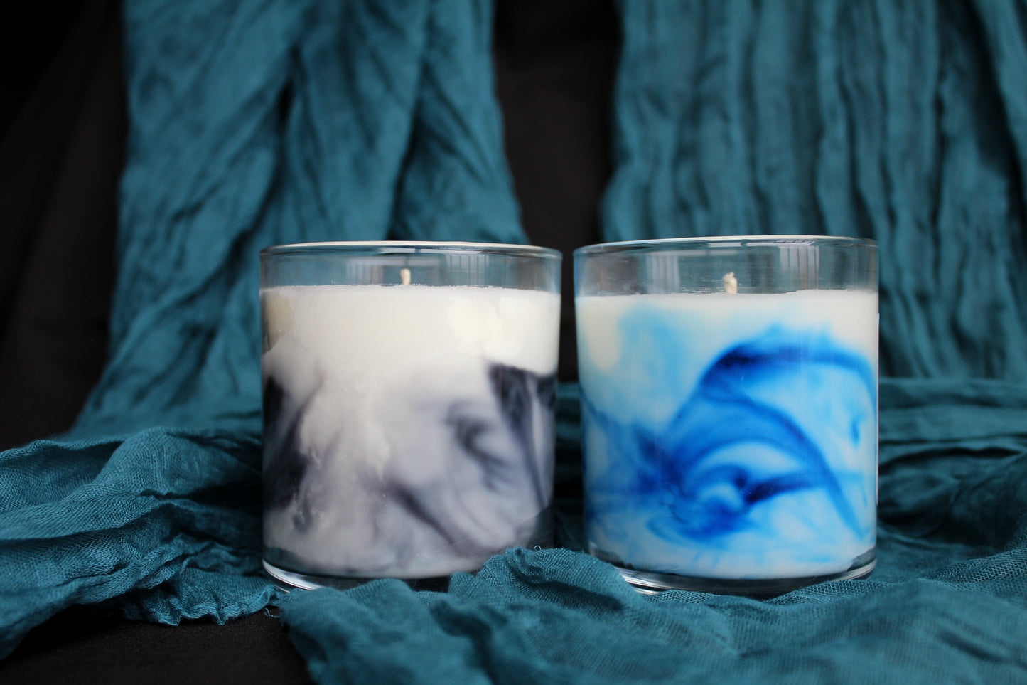 Two candles with swirling designs in glass containers on a textured teal fabric background.