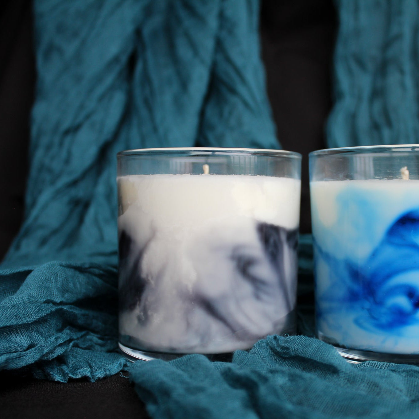 Two candles with swirling designs in glass holders on a textured teal fabric background