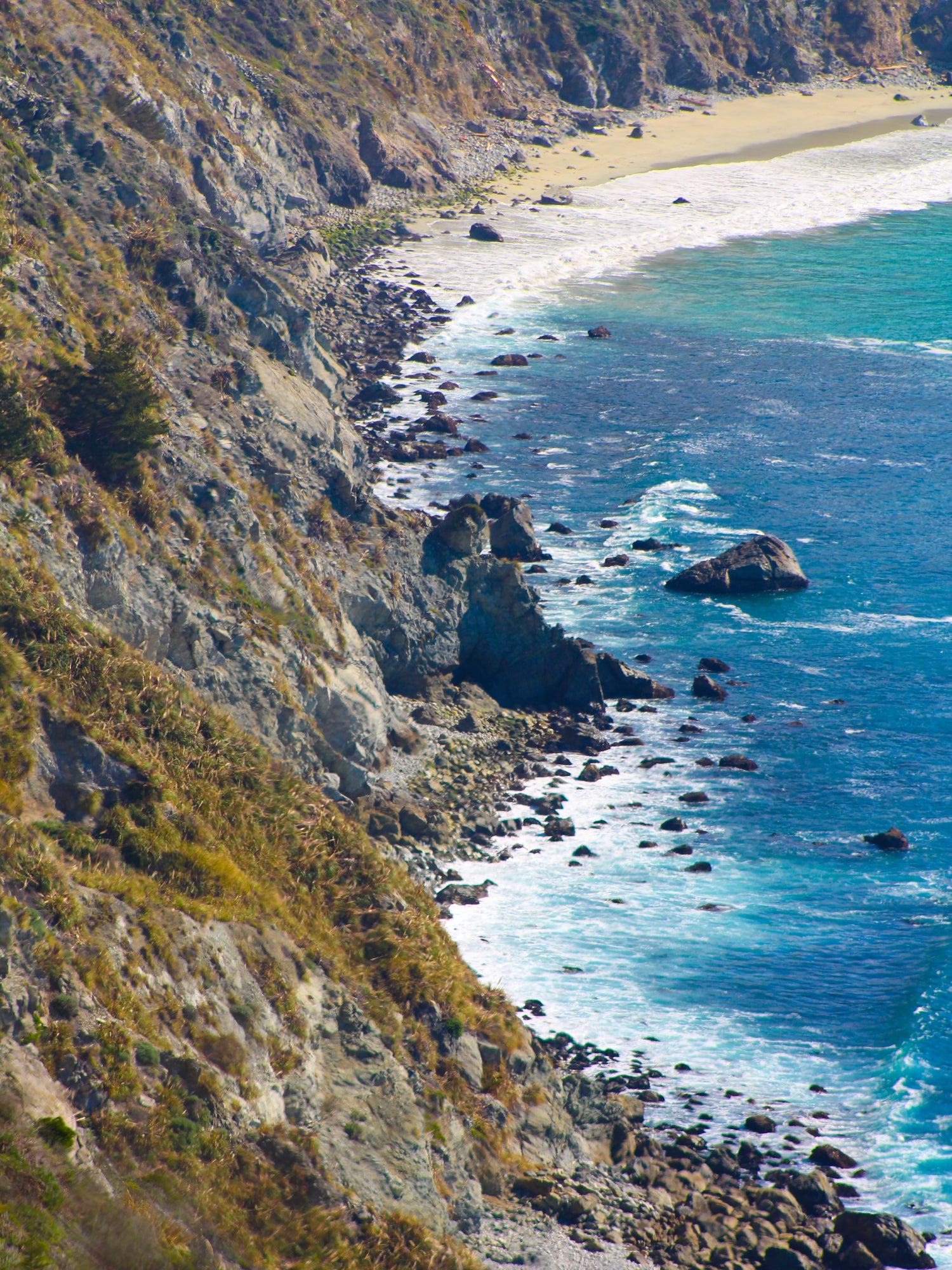 Big Sur coastline, waters are shades of blue and the waves are crashing white along a mossy hillside.