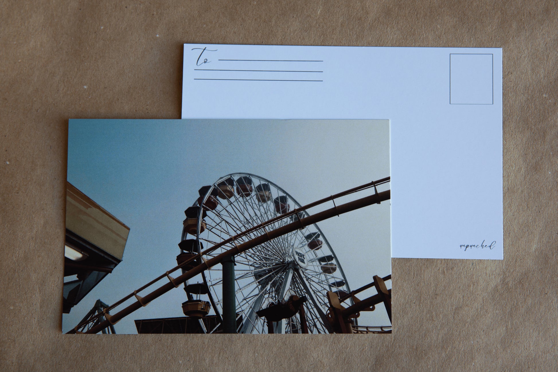 Photo of a Ferris Wheel in warm tones. Colors of the Ferris Wheel seats/ buckets are red, blue and yellow. In the foreground there is a rollercoaster track that is yellow with green posts. There are other metal posts pictured that aren't as decipherable.Back of the post card with "To" and stamp square. Logo in the bottom right corner.