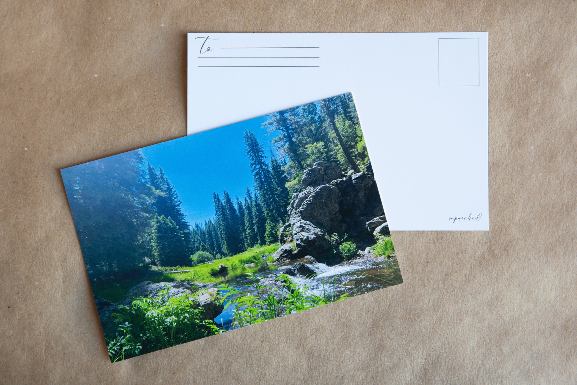 Back of the post card with "To" and stamp square. Logo in the bottom right corner. Cascading river flow on rocks, in the middle of a coniferous forest. Vibrant green shrubbery with tall fir and pine trees surrounding a grassy valley. Boulders on the right side of the picture, just behind the river flowing. 