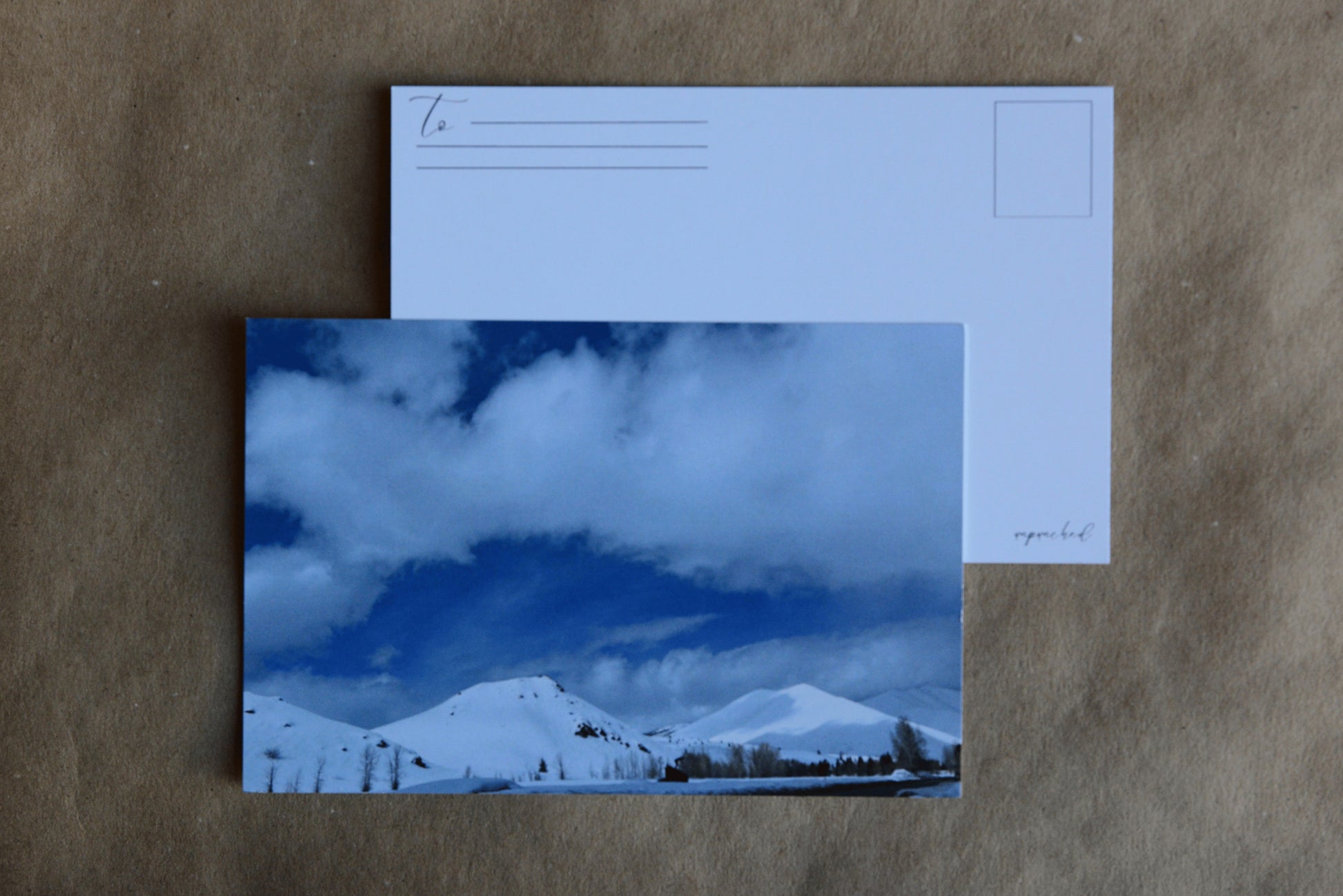 Back of the post card with "To" and stamp square. Logo in the bottom right corner. Mountains covered in snow in Sun Valley, Idaho. The road with ice walls in the foreground. The sky is blue with clouds that are faint when compared to the snow covered mountains with the sun hitting them, creating shadows on the furthest mountain.