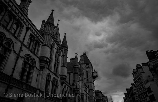 Black and white photo of a street view of a building in London. The clouds are rolling. There is a clock on the side of one of the building in the foreground.