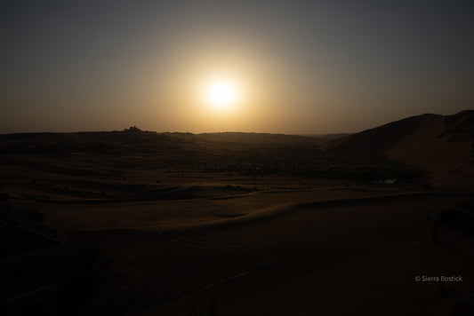 Sunset over a desert landscape with silhouettes of dunes and structures.