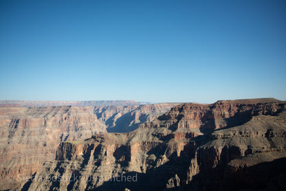 Wide span photo of the grand canyon. The left side of the photo, bright exposure of the canyon rock. The left side of the photo is shadowed canyon rock. The background is an ombré blue dark to light, top to bottom, respectively. Watermarked.