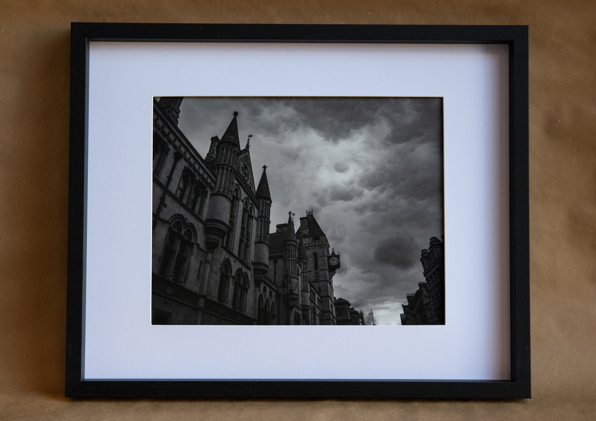 Black and white photo of a street view of a building in London. The clouds are rolling. There is a clock on the side of one of the building in the foreground. White matting of an 11x14 with a black frame.