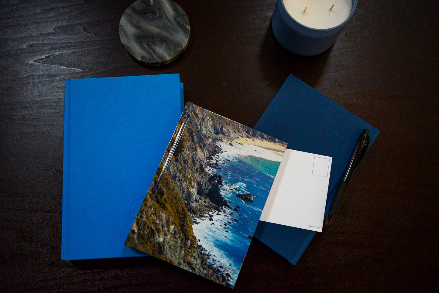 Diffraction hardcover notebook against the blue covered books. Candle, coaster and pen in the picture. Along with unpunched postcard