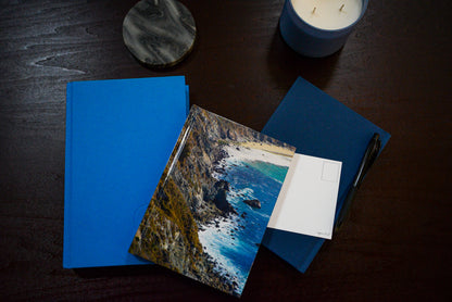 Diffraction hardcover notebook against the blue covered books. Candle, coaster and pen in the picture. Along with unpunched postcard