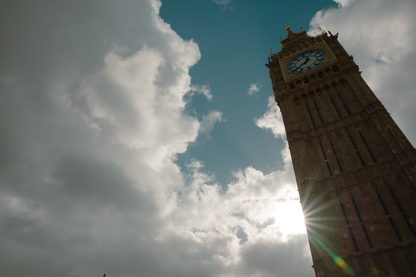 Elizabeth's Tower in the right corner of the frame. Tan with gold accents around Big Ben (clock) which is a cream color with a blue around the clock face. Black hour markings with black hands. The backgrouns is a warm blue sky with many clouds. The sun is peaking through the bright white clouds beaming off the side of the tower creating a rainbow colored ray in the foreground. 