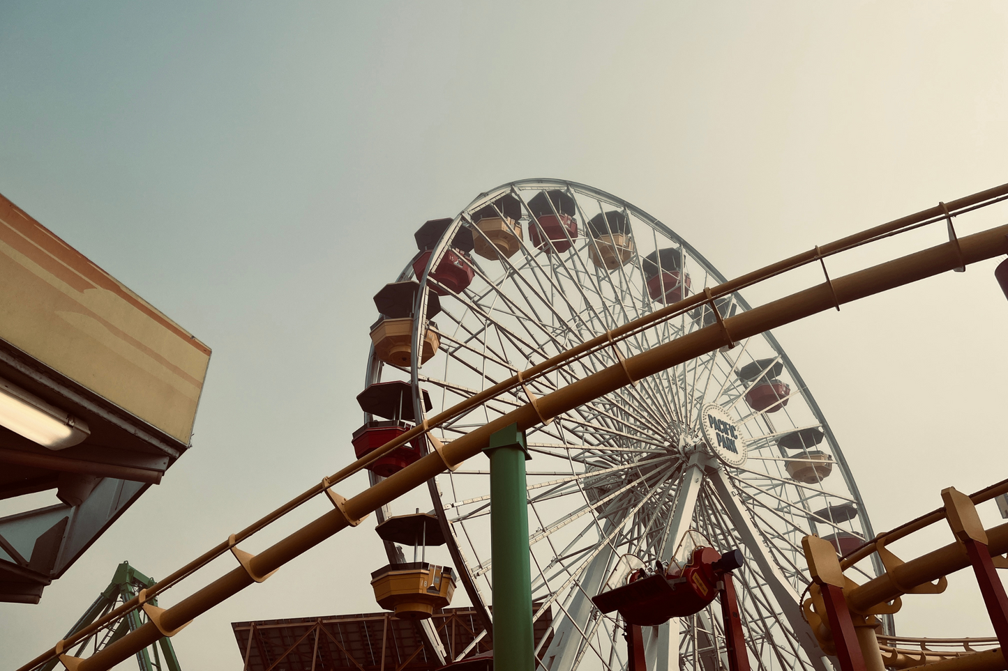 Photo of a Ferris Wheel in warm tones. Colors of the Ferris Wheel seats/ buckets are red, blue and yellow. In the foreground there is a rollercoaster track that is yellow with green posts. There are other metal posts pictured that aren't as decipherable. 