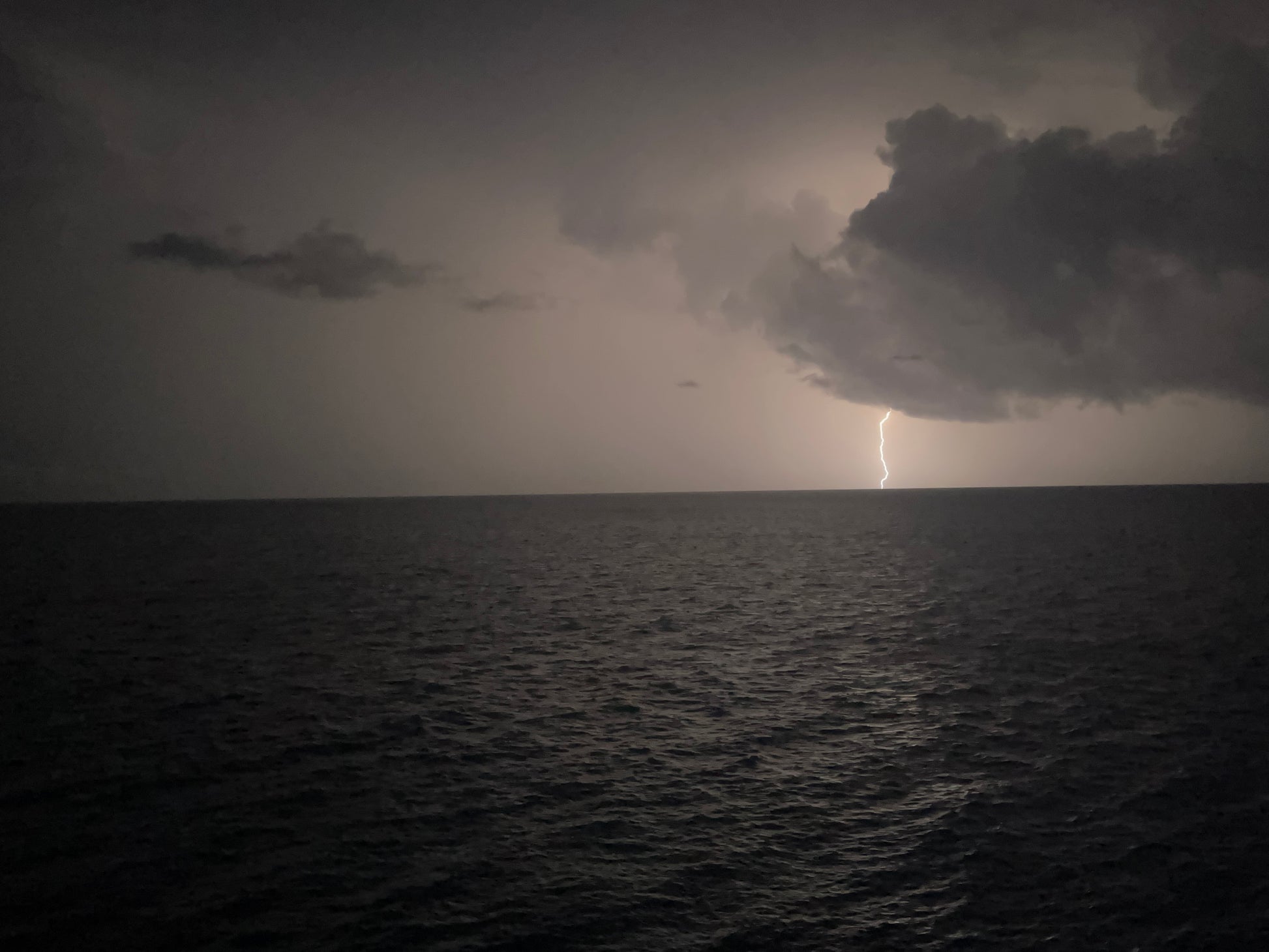 Black and white picture of a thunderstorm in the Bahamas. One cloud with a lightning striking the water. The peaks of the waves are illuminated by the lighting. 