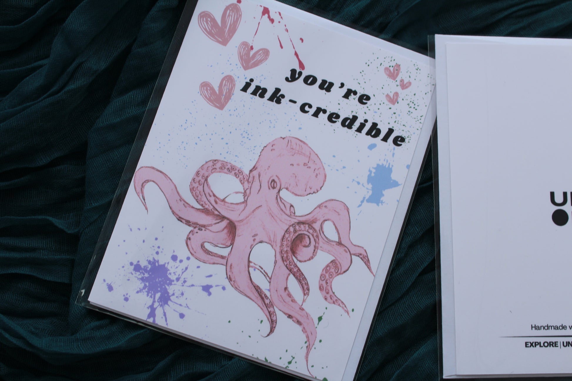 packaged ink-credible card with octopus and paint splatter on the front, on teal linen background 