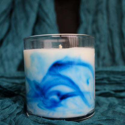 Candle with blue and white swirls in a glass container on a textured green surface