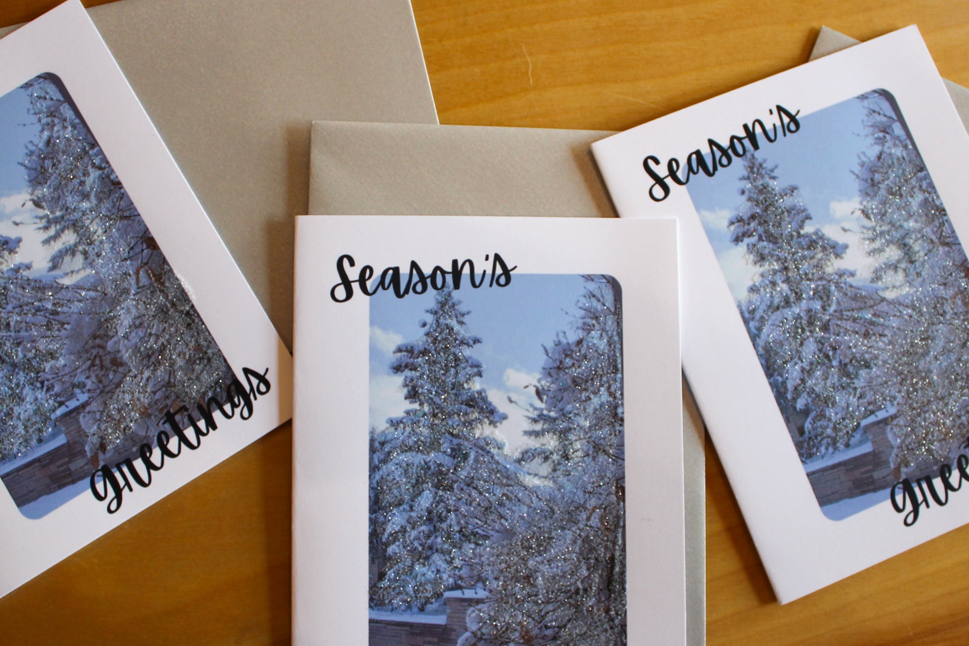 Three greeting cards with snowy tree designs and 'Season's Greetings' text on a wooden surface.