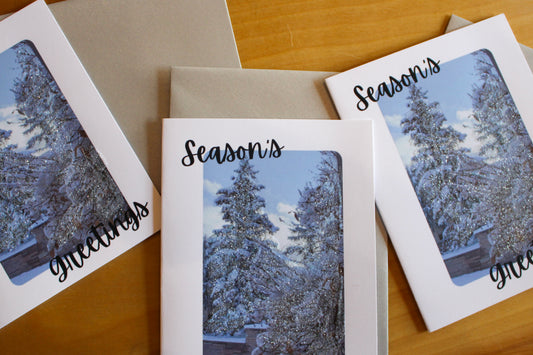 Three greeting cards with snowy tree designs and 'Season's Greetings' text on a wooden surface.