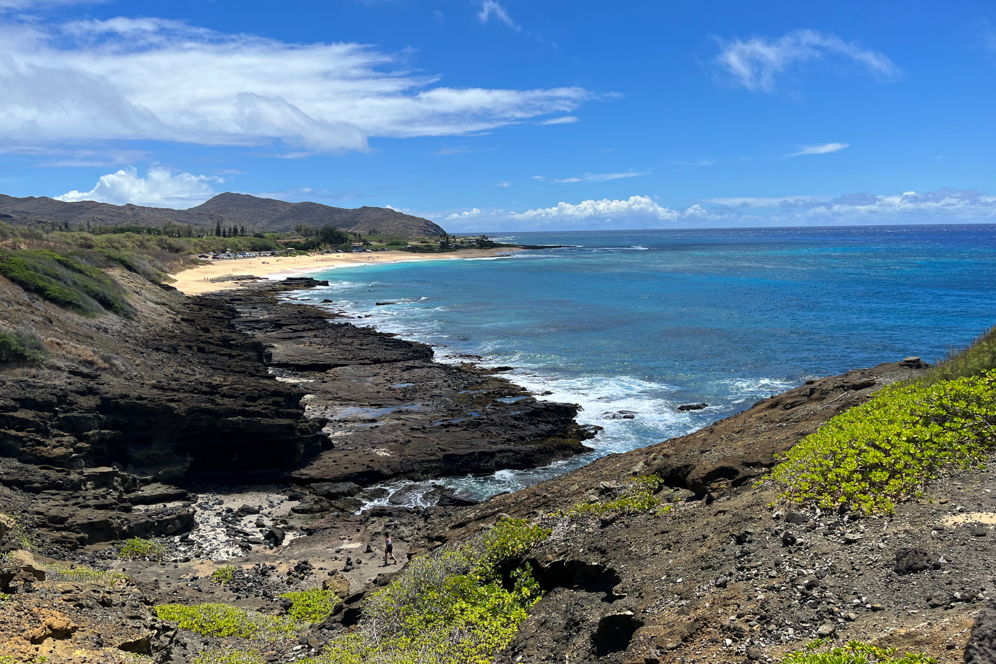 Wide/high view of Sandy's Beach in Oahu, Hawai'i. In the foreground there is mountain with erosion and grassy patches. In the middle ground there is a significant amount of jet (black stone) and a person walking towards it. In the background there is Sandy's Beach commonly known as Breakneck Beach, with a mountain and point in the further distance. The water is blue, with the white tide crashing the jet. 