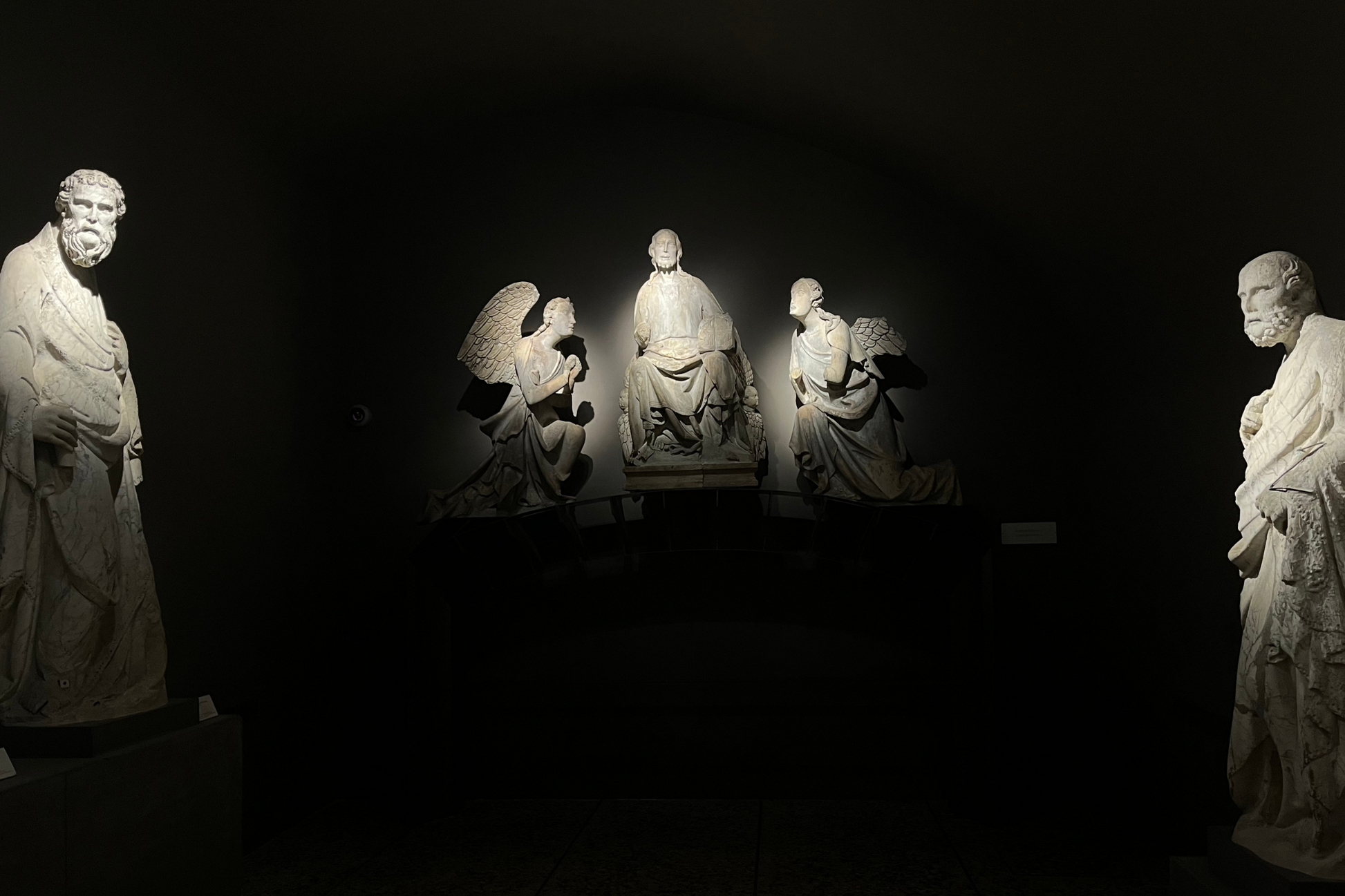 Two religious statues standing in the foreground. In the background there is a figure sitting and he is surrounded by two kneeling angels looking at him. The statues are white with the surrounding areas being black with a spotlight illuminating the statues. 