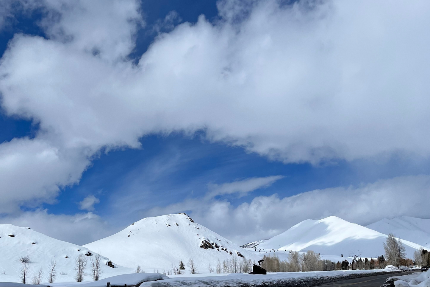 Mountains covered in snow in Sun Valley, Idaho. The road with ice walls in the foreground. The sky is blue with clouds that are faint when compared to the snow covered mountains with the sun hitting them, creating shadows on the furthest mountain. 