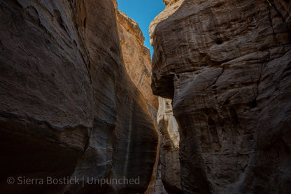 Upward view of the Al Siq Canyon in Jordan. Surrounded by striated red rock, that looks gold. At the top center of the picture there is a clear blue path of sky.