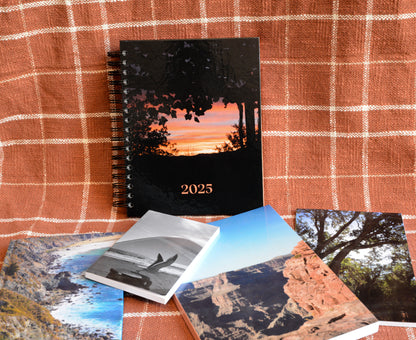 peach and white plaid background. 2025 spiral planner standing. those who wander collection laying down (4 notebooks) diffraction (big sur coast), take off (bird taking off of a trash can), rugged (grand canyon), deciduous (trees).