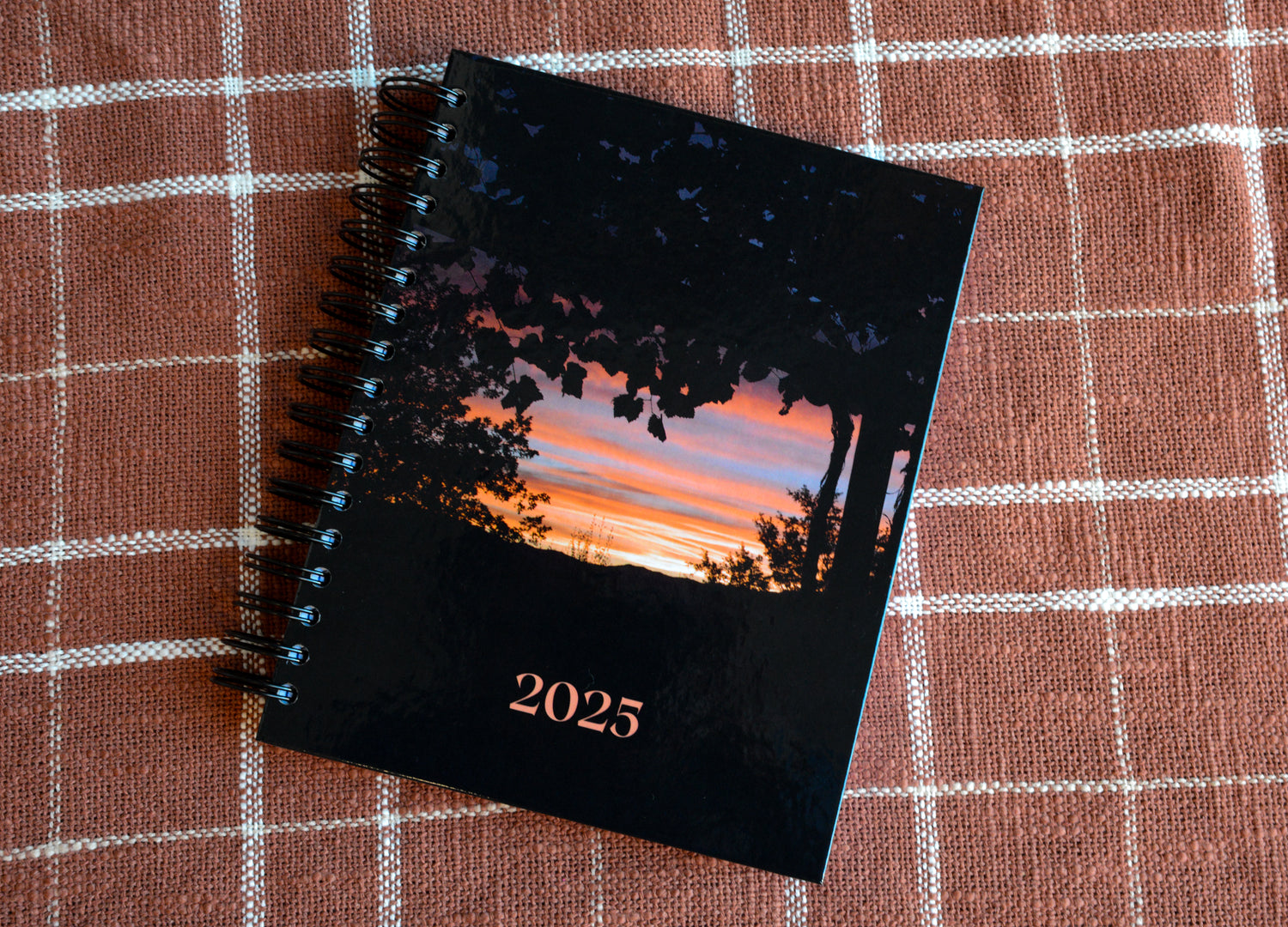 2025 Weekly Planner, black spiral hardcover with vines and shrubs shadowed black. blue, peach, and orange sunset peeking through the black. on peach plaid white background